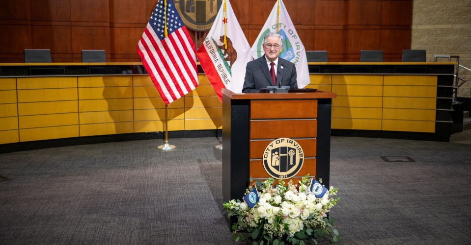 Mayor Larry Agran standing at a podium with the American flag, California flag, and City of Irvine flag behind him. 