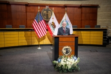 Mayor Larry Agran standing at a podium with the American flag, California flag, and City of Irvine flag behind him. 