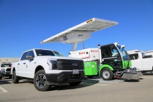 A white electric truck and green and white street sweeper are parked under a solar-powered EV charger.